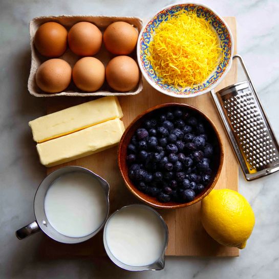 Buttermilk Blueberry Pancake Casserole Recipe 4 The image shows a wooden surface with various baking ingredients arranged neatly. There is a box of brown eggs with smooth shells in the top left, two rectangular sticks of pale butter below the eggs, and a small pile of bright yellow lemon zest nearby. A brown bowl filled with fresh dark blueberries is at the bottom left. Two metal measuring cups contain white liquid, likely milk or cream, positioned near the center; one cup is larger than the other. A bright yellow lemon sits at the bottom right. A metal grater lies at the top right, with a colorful cup partially visible behind it, all on a white marbled surface background. Photo taken with an iphone --ar 4:5 --v 7