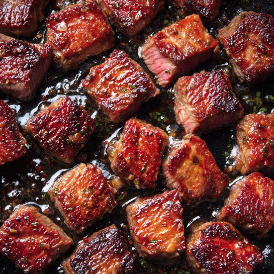 The image shows many small, square pieces of cooked steak in a black pan. Each piece has a crispy, dark brown outside with visible char marks and a juicy, pinkish center. The meat is glistening with a light layer of oil, making it look tender and flavorful. The pieces are spread evenly, covering the pan’s surface with some pieces slightly overlapping. The black pan contrasts with the rich brown and pink colors of the steak. photo taken with an iphone --ar 4:5 --v 7