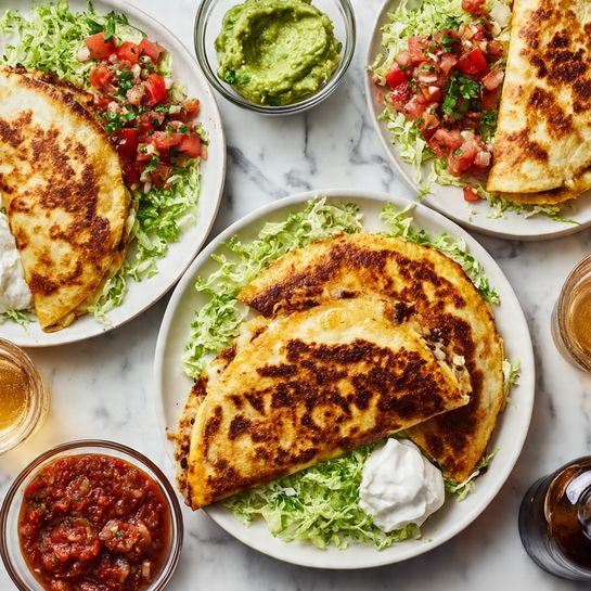 The image shows three white plates on a white marbled surface, each holding three golden-brown quesadillas that are folded in half. The quesadillas have a slightly crispy texture with darker spots and are placed on a bed of shredded green lettuce. Each plate has different toppings: one with diced red tomatoes and onions mixed with herbs, another with the same topping plus a dollop of green guacamole and white sour cream, and the third with diced tomatoes and a small amount of white sauce on the side. Around the plates are clear bowls filled with chunky red tomato salsa and creamy green guacamole. A brown bottle and a glass of light beer are also visible on the white marbled surface. photo taken with an iphone --ar 4:5 --v 7