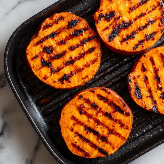 Four thick slices of bright orange sweet potato arranged on a black grill pan, each slice showing dark, straight grill lines across the top surface. The sweet potatoes have a slightly shiny texture with some areas showing deeper char marks, especially on the edges. The grill pan has ridges and a glossy, slightly oily finish. The image is focused closely, filling the frame with the sweet potatoes and grill details, all placed on a white marbled surface photo taken with an iphone --ar 4:5 --v 7