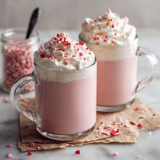 Two clear glass mugs with handles hold a pink drink filling most of each mug as the bottom layer. On top, a thick white whipped cream layer covers each mug, sprinkled with small red, white, and pink heart-shaped sprinkles. The mugs rest on pieces of crumpled brown paper placed on a white marbled surface, with some heart sprinkles scattered around. In the background, slightly out of focus, is a clear jar with a spoon inside. Photo taken with an iphone --ar 4:5 --v 7