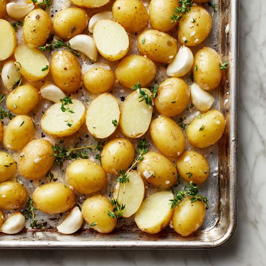 A baking tray filled with small, halved yellow potatoes scattered all over, some showing the smooth pale yellow inside and others the slightly shiny golden skin outside with light brown spots. Sprigs of green thyme and whole garlic cloves with creamy white skin are spread among the potatoes, along with small grains of salt and black pepper. The tray surface reflects a faint light, creating a subtle shine on the potatoes. The scene is on a white marbled texture. photo taken with an iphone --ar 4:5 --v 7