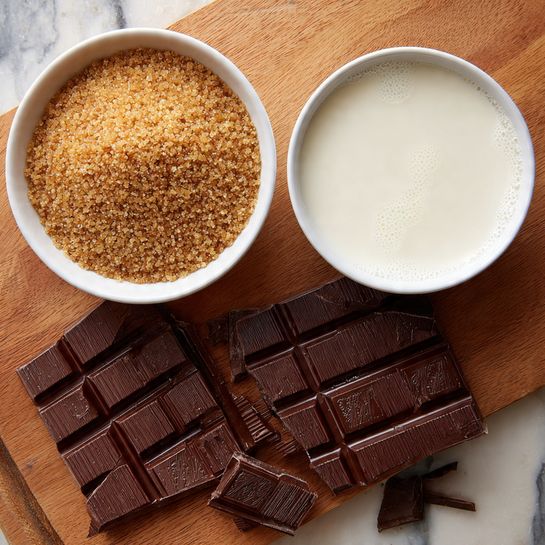 The image shows two white bowls placed on a wooden surface, one filled with granulated brown sugar showing a rough, grainy texture, and the other filled with smooth white milk with small bubbles floating on top. Beside the bowls, there is an open dark chocolate bar on its wrapper with distinct rectangular segments, some pieces broken off and placed near the bar. The chocolate pieces have a glossy finish and embossed designs. The scene has a warm, natural feel with a white marbled texture background added. Photo taken with an iphone --ar 4:5 --v 7