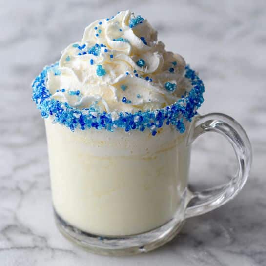 The image shows a clear glass mug filled with a creamy white drink. The rim of the mug is decorated with bright blue sugar crystals that add a colorful touch. On top of the drink, there is a large swirl of white whipped cream sprinkled with small blue sugar bits that match the rim. The mug has a clear handle on the right side and is placed on a surface with a white marbled texture. The background is softly blurred, keeping focus on the drink. Photo taken with an iphone --ar 4:5 --v 7