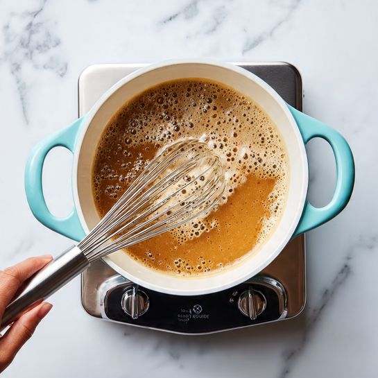 A top view of a small white pot with light blue handles filled with a bubbling light brown liquid being stirred with a metal whisk that has a black handle. A woman's hand holds the whisk from the left side. The pot sits on a stainless steel countertop burner with black edges and control knob, all placed on a white marbled surface. Photo taken with an iphone --ar 4:5 --v 7