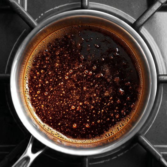 Pineapple Upside Down Cake Recipe 4 A close-up top view of a stainless steel saucepan on a black stove cooktop, showing a thick dark brown sauce boiling with small bubbles and a slightly uneven texture inside the pan. The shiny metal rim contrasts with the rich, glossy sauce inside. The background is the black stove surface with white circular markings. photo taken with an iphone --ar 4:5 --v 7