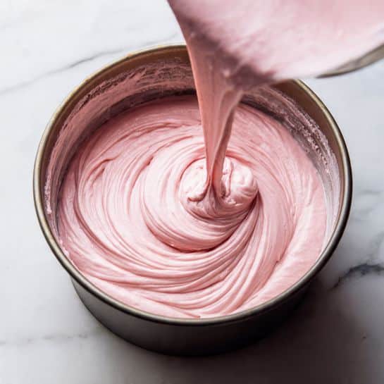 A thick, smooth, light pink batter is being poured in soft folds into a round, dark metal baking pan. The batter forms several layered swirls at the base, showing its creamy and slightly dense texture. The pan sits on a surface with a white marbled texture, creating a clean and bright backing for the soft pink batter inside. photo taken with an iphone --ar 4:5 --v 7