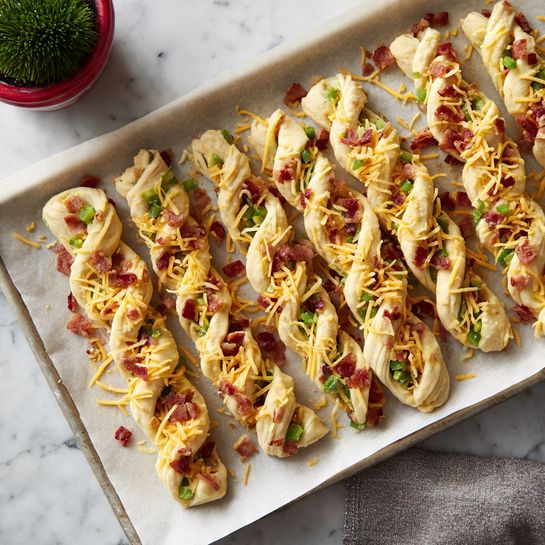 The image shows several twisted pastry sticks lined up on a baking sheet covered with parchment paper, placed on a white marbled surface. Each pastry stick has a light yellow dough base filled with small pieces of green jalapeño slices, bits of red bacon, and sprinkled generously with shredded orange cheddar cheese on top. The pastries appear raw, ready to be baked, with the cheese and jalapeños scattered unevenly but covering each piece well. In the top left corner, there's a glimpse of a green plant in a red and white pot, and the bottom part of the image shows a person's legs and gray pants. Photo taken with an iphone --ar 4:5 --v 7