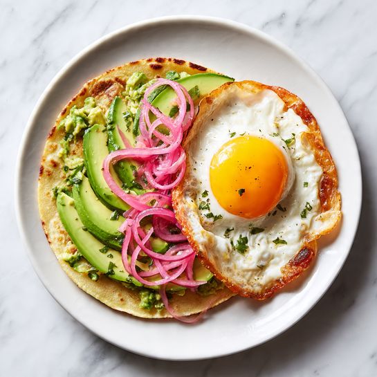 The image shows two stages of a tortilla dish on a white plate with a white marbled textured background. On the left side, a single tortilla layer is topped with thin slices of avocado, evenly spread and creating a smooth, green layer with soft texture and some chopped pieces visible. On the right side, the same tortilla with avocado now has a fried egg placed on top, the egg white is cooked with slightly crispy brown edges, and the bright yellow yolk is whole and shiny. There are thin, curly slices of pink pickled onions artfully placed on the egg. The dish has a fresh and colorful look with green, white, yellow, and pink tones, and the textures vary from soft avocado to crispy egg edges. photo taken with an iphone --ar 4:5 --v 7