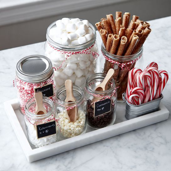 A white tray holds several clear glass jars and metal containers filled with different small treats, all arranged neatly on a white marbled surface. The largest jar is full of white mini marshmallows and has a red and white string tied around its neck. Next to it stands a jar filled with rolled wafer sticks, light brown with darker stripes, also tied with red and white string. In front, there are jars with small wooden scoops inside, holding white chocolate chips, dark chocolate chips, toffee bits, and crushed white candy pieces. Another glass jar contains small red and white striped candy canes, and a metal container to the side has individually wrapped red and white peppermint sticks. Each jar has a small black chalkboard label with white writing, and the whole setup sits on a rectangular white tray. Photo taken with an iphone --ar 4:5 --v 7