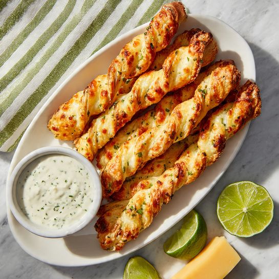 The image shows crispy twisted cheese sticks with bits of green herbs and small pieces of bacon, golden brown and flaky in texture. Several sticks are in a clear glass jar in the background, while one stick is held by a woman's hand, dipped halfway into a smooth, creamy white sauce with green herbs in a small white bowl. The bowl sits on a dark green plate, and more cheese sticks rest on the plate beside the bowl. The setting is bright with sunlight, with a white marbled surface under everything and some green plants to the side. Photo taken with an iphone --ar 4:5 --v 7