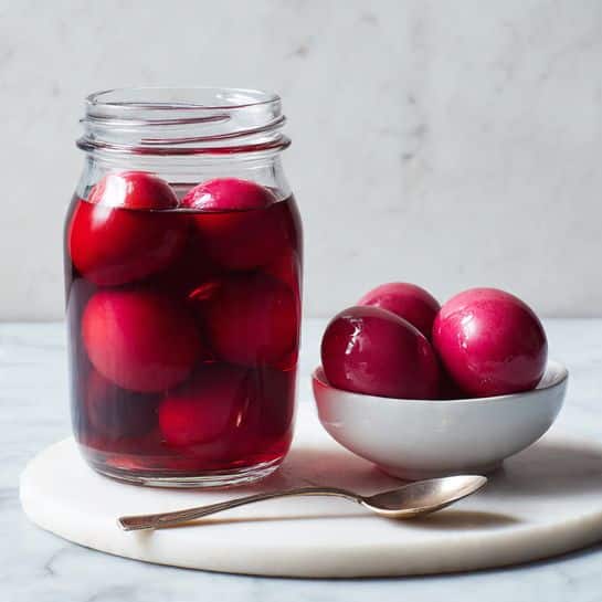 The image shows a clear glass jar filled with deep red liquid, soaking whole eggs that have taken on a vibrant pinkish-red color. The jar is placed on a round white marble board. Next to the jar, there is a small white bowl holding two more pinkish-red eggs with a smooth, shiny surface. A tarnished silver spoon lies in front of the jar on the white marble board. The background is simple with a light wall and white marbled texture beneath. photo taken with an iphone --ar 4:5 --v 7