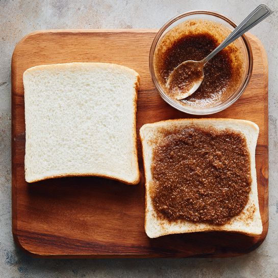 The image shows two square pieces of white sandwich bread placed flat on a wooden surface, the left slice plain white and the right slice evenly spread with a thick layer of dark brown, grainy textured spread. Above the bread slices is a small clear glass bowl containing more of the dark brown spread, with a silver spoon resting inside it. The background is a wooden cutting board. photo taken with an iphone --ar 4:5 --v 7