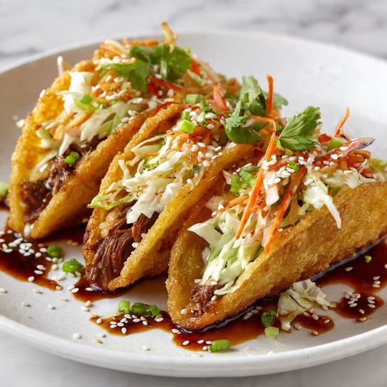 A white plate holds five golden brown fried dumplings arranged in a row. Each dumpling is filled with shredded light brown meat and topped with thinly sliced white and purple cabbage. Bright green chopped herbs and scallions are sprinkled on top along with white sesame seeds. A reddish-brown sauce is drizzled over the dumplings and plate unevenly. The plate rests on a white marbled surface. photo taken with an iphone --ar 4:5 --v 7
