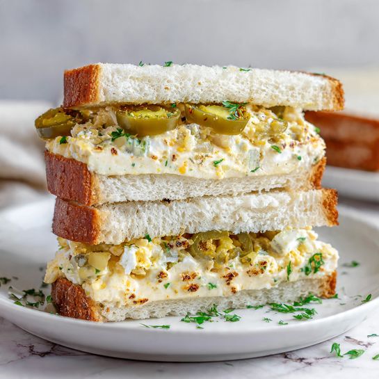 The image shows two halves of a sandwich stacked on top of each other on a white plate, placed on a white marbled surface. Each half has three visible layers: the bottom and top layers are thick slices of white bread with a slightly browned crust, and the middle layer is a creamy mixture with a mix of orange cheese, white chopped egg pieces, small green herbs, and slices of green jalapeño peppers. The cheese topping shows some browned spots, giving a slightly toasted look, and the green jalapeños add a fresh contrast in color and texture. Small chopped herbs are also visible scattered on the plate around the sandwich. photo taken with an iphone --ar 4:5 --v 7
