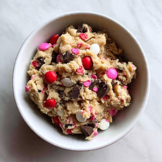 A white bowl filled with cookie dough that has a light tan color and a rough texture. Mixed throughout the dough are pieces of dark chocolate cookies and colorful round candy pieces in red, white, and pink. The dough looks thick and chunky, sitting on a white marbled surface. photo taken with an iphone --ar 4:5 --v 7
