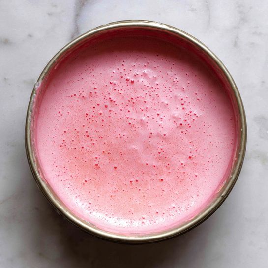 A round silver bowl filled with a smooth, thick pink batter that has small bubbles on the surface. The batter evenly coats the inside edges of the bowl, leaving a slight thicker line along the side. The background is a white marbled surface that brightens the image. photo taken with an iphone --ar 4:5 --v 7