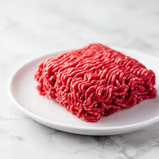 A close-up image of a single block of raw ground meat placed in the center of a white plate. The meat is a deep red color with a granular texture, showing tightly packed strands and small bits of white fat mixed inside. The plate is set on a white marbled surface, giving a clean and simple background. The lighting highlights the meat’s shiny, fresh appearance. photo taken with an iphone --ar 4:5 --v 7
