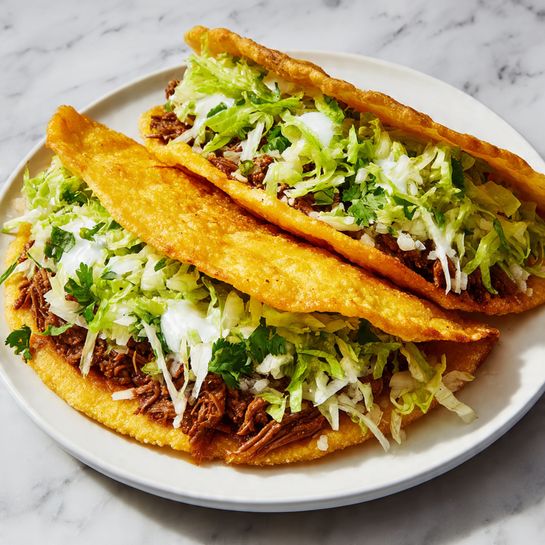 The dish shows two thick golden brown fried tortillas slightly puffed, acting as the top and bottom layers of each taco-like sandwich. Inside, there is a layer of shredded green lettuce mixed with white sharp-looking shredded cheese scattered across the top. Beneath the greens, tender pieces of brown cooked meat fill the middle. The sandwiches are served on a round white plate, which sits on a white marbled surface. Photo taken with an iphone --ar 4:5 --v 7
