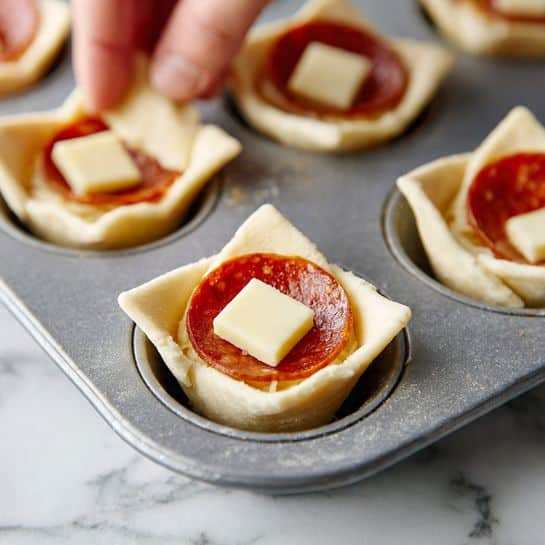 The image shows a close-up of a gray muffin tray with several cups filled with dough, each cup lined with a triangle-shaped layer of pale, soft dough with uneven edges. Inside the dough, there are two round, reddish-orange slices of pepperoni resting flat, topped with a small rectangular piece of pale yellow cheese at the center, creating a layered effect. The tray is placed on a white marbled surface, and the focus is on one cup in particular, with parts of a woman's hand visible holding a piece of dough in the background. photo taken with an iphone --ar 4:5 --v 7