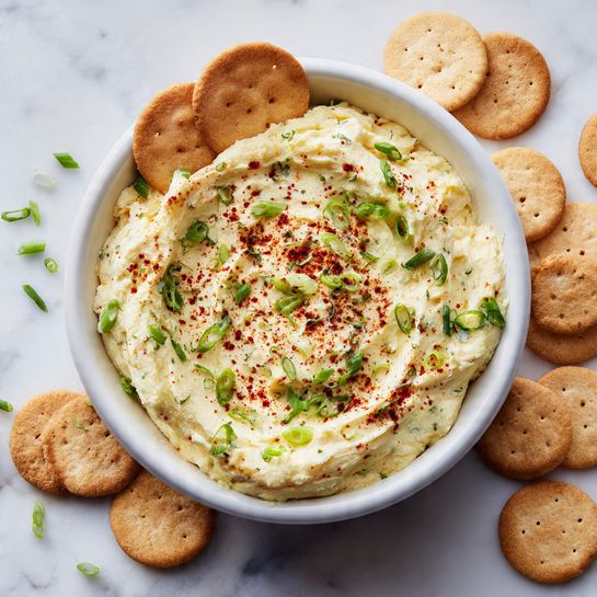 A white bowl filled with a creamy, light yellow spread is placed on a white marbled surface, surrounded by round beige crackers. The spread has a smooth yet slightly fluffy texture with a small well in the center sprinkled with reddish-brown seasoning and finely chopped green herbs scattered on top. One round cracker is partially dipped into the spread, resting at the edge of the bowl. In the background, there's a blurred bowl with green herbs and some out-of-focus colorful fruits. Photo taken with an iphone --ar 4:5 --v 7