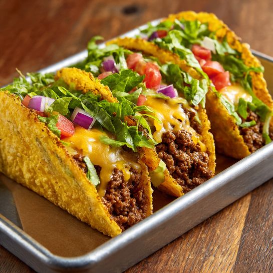 The image shows three tacos in a silver tray on a wooden surface. Each taco has a crispy, golden-yellow shell that looks slightly browned and bubbly. Inside, there is a layer of cooked ground meat with a rich brown color. Over the meat, melted light yellow cheese is spread, blending with the meat below. Fresh green lettuce pieces and small red tomato chunks sit on top, adding bright color. Small bits of purple onion are scattered above the lettuce and tomato, giving a fresh contrast. The tacos are closely placed, filling the tray fully. Photo taken with an iphone --ar 4:5 --v 7