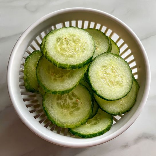 Thin, round slices of cucumber with dark green edges and light green centers are stacked loosely inside a white plastic bowl with vertical slits all around. The cucumber slices show a moist texture with small seeds visible, and some are slightly overlapped, creating a layered look. The background surface is a white marbled texture. photo taken with an iphone --ar 4:5 --v 7