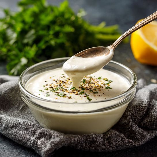A glass bowl filled with a smooth, creamy white sauce with some black pepper sprinkled on top. A silver spoon is dipped into the sauce, lifting a small spoonful. In the background, there is a green parsley bunch and a bright yellow lemon wedge. The bowl is placed on a dark surface, with a gray textured cloth near it. The overall scene has natural lighting and a cozy feel. photo taken with an iphone --ar 4:5 --v 7