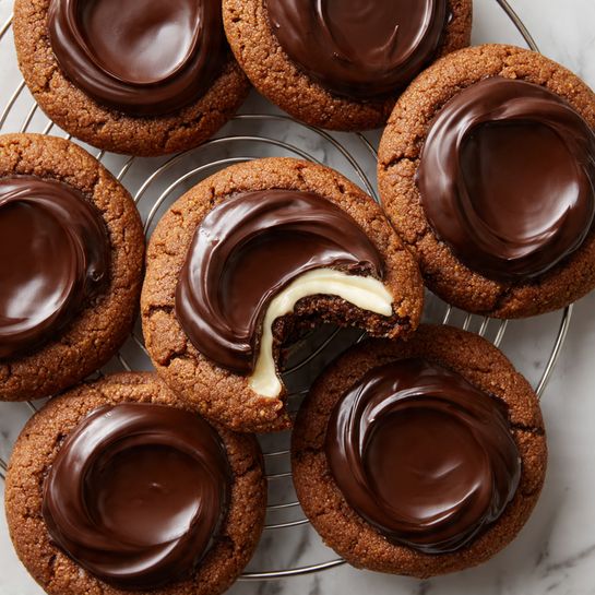 The image shows a stack of three round chocolate cookies on a white marbled surface, each cookie topped with smooth, glossy dark chocolate. The top cookie is broken in half and placed on top of the second cookie, revealing a thick, white marshmallow filling inside. The broken cookie pieces show a soft, crumbly texture on the chocolate cookie with the marshmallow creamy and fluffy within. The background is blurred with other cookies scattered, keeping the focus on the stacked cookies in the center. photo taken with an iphone --ar 4:5 --v 7