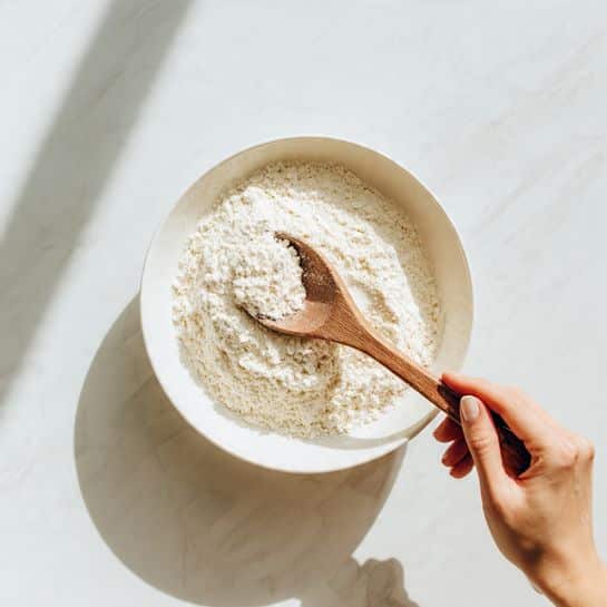A white bowl filled with a layer of white flour is placed on a white marbled surface. A wooden spoon is scooping some of the flour, held by a woman's hand entering from the bottom right corner of the image. The scene is bright and minimal with soft natural light. Photo taken with an iphone --ar 4:5 --v 7