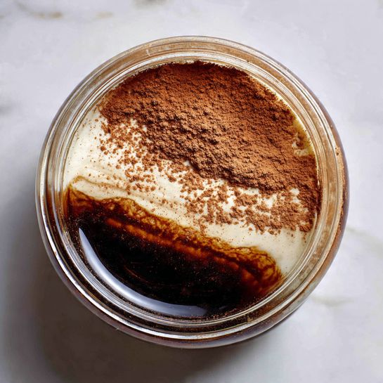 A clear glass jar filled with three visible layers seen from above: the bottom layer is dark and shiny, resembling a thick syrup or chocolate sauce, the middle layer is a creamy white substance with a smooth texture, and the top layer is a light brown cocoa powder dusted unevenly, partially covering the white layer below. The jar sits on a white marbled surface. Photo taken with an iphone --ar 4:5 --v 7