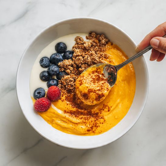 A white bowl sits on a white marbled surface, filled with three main layers: the bottom layer is a smooth, white yogurt with a glossy texture, partially covered by a thick, bright orange pumpkin puree layer that has a soft, slightly chunky texture sprinkled lightly with brown spices on top. Scattered around the pumpkin puree are fresh blueberries and a deep red raspberry, along with some crunchy, golden granola clusters adding texture contrast. A woman's hand holds a spoon above the bowl, lifting a scoop of the bright orange pumpkin puree topped with a sprinkle of brown spices. photo taken with an iphone --ar 4:5 --v 7