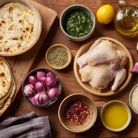 The image shows three raw chicken pieces placed neatly on a wooden board with a small knife beside them on the left side. Below the board, there is a stack of flatbreads with a light brown color and dark spots on top, sitting on a white marbled surface. In the bottom middle, a white bowl contains a creamy white sauce with green herbs mixed in. Around the board and bowl, there are various small containers holding spices and oils, with colors ranging from light yellow to dark red. To the right, a cluster of fresh green dill and a halved head of pale green lettuce are placed close together. On the bottom right, a white bowl is filled with sliced cucumber rounds and whole small cucumbers. Several lemon halves and whole lemons are scattered on the marbled surface, along with a bulb of garlic near the lower right corner. photo taken with an iphone --ar 4:5 --v 7