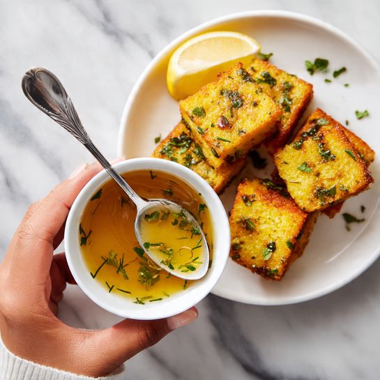 A woman's hand holds a small white bowl filled with a light golden sauce containing small green herb leaves, with a silver spoon resting in the bowl. In the background, there is a white plate filled with several golden brown, rectangular pieces of cooked food garnished with small green herbs, and a lemon wedge is placed on the edge of the plate. The scene is set on a white marbled surface. photo taken with an iphone --ar 4:5 --v 7