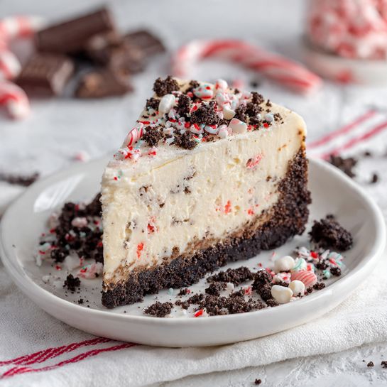 A single slice of cheesecake sits on a white plate, showing two distinct layers: a thick, creamy pale layer with small red and white pieces mixed inside, and a dark crumbly base that looks like crushed chocolate cookies. The top of the cheesecake slice is smooth and white with scattered small crushed red and white candy pieces and cookie crumbs. Around the plate, there are extra cookie crumbs and candy bits. The plate is placed on a white cloth with red stripes, and the background features a white marbled texture with some candy canes and chocolate pieces slightly blurred. Photo taken with an iphone --ar 4:5 --v 7