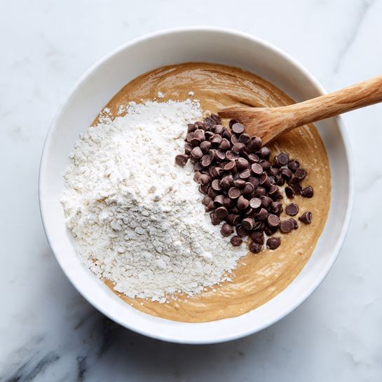 A white bowl sits on a white marbled surface, filled with three layers of ingredients for baking. The bottom layer is a smooth, light brown batter that covers the base and part of the side. On top of that, there is a thick layer of white flour mostly covering one half of the bowl. Piled on the flour is a cluster of small, dark brown chocolate chips. A wooden spoon with a light color rests inside the bowl, partly on the flour and batter mixture. photo taken with an iphone --ar 4:5 --v 7