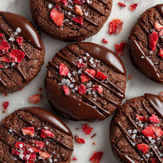 The image shows several round, dark brown chocolate cookies with a rough, textured surface. Each cookie has small pieces of bright red strawberry bits embedded and scattered on top, along with a light sprinkle of white sea salt flakes. Some cookies have a smooth, glossy dark chocolate coating on the bottom layer, while the top layer has thin drizzles of dark chocolate all over. The cookies are arranged closely on a white marbled surface, with some chocolate drizzles and crumbs around them. Photo taken with an iphone --ar 4:5 --v 7