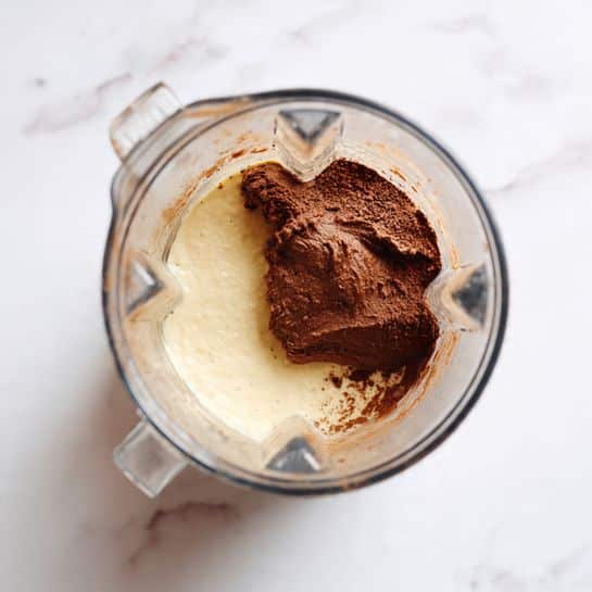 A clear blender jar with two main layers inside is shown from above. The bottom layer is a light cream color and looks smooth and creamy, filling roughly half the jar. On top of this, there is a large dollop of thick brown paste, sitting mostly on one side of the creamy base with some paste slightly touching the jar wall. The background is a white marbled surface. photo taken with an iphone --ar 4:5 --v 7