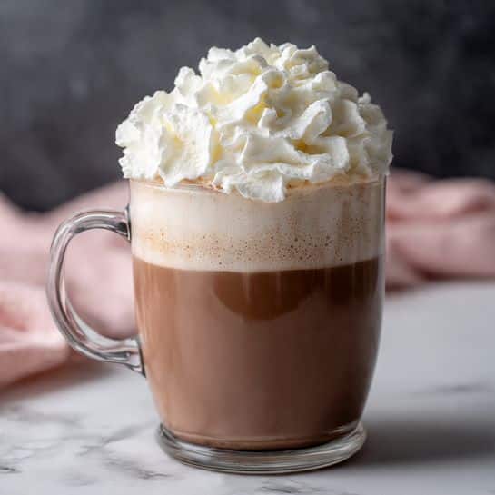 A clear glass mug filled with three layers: the bottom layer is smooth, dark brown hot chocolate; the middle layer shows some foam and slight melting around the edges creating a soft gradient from darker to lighter brown; the top layer is a large, fluffy swirl of bright white whipped cream, piled high with textured peaks. The mug handle is visible on the left side, and the mug sits on a white marbled surface with a blurred dark background, part of a pink cloth is seen on the left side near the mug. Photo taken with an iphone --ar 4:5 --v 7