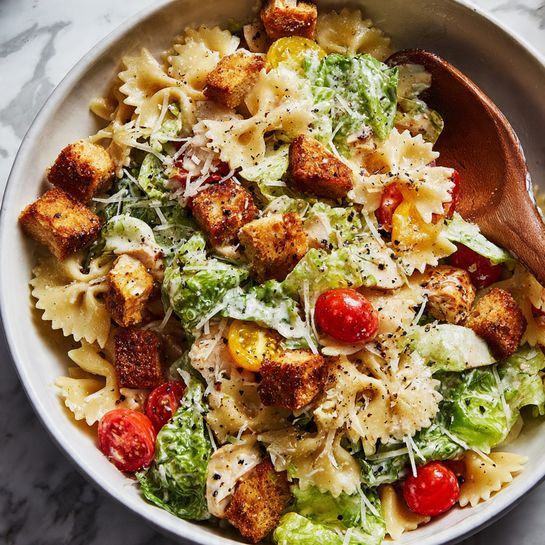 A close-up view of a white bowl filled with a layered pasta salad on a white marbled surface, the bottom layer made of cream-colored farfalle pasta. Scattered throughout the pasta are bright green Romaine lettuce leaves and halved red cherry tomatoes. The salad is topped with well-seasoned, golden-brown croutons and lightly browned, diced pieces of cooked chicken. A creamy dressing coats the ingredients, with a sprinkle of shredded white cheese and cracked black pepper on top. A wooden spoon rests inside the bowl on the right side. Photo taken with an iphone --ar 4:5 --v 7