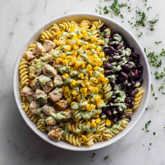 The image shows a close-up of a colorful pasta salad on a wooden spoon above a larger bowl. The salad has three main layers visible: creamy, light beige rotini pasta with slightly glossy texture, pieces of white chicken breast with a soft fibrous look placed on top and around the pasta, yellow corn kernels scattered throughout, black beans mixed in for contrast, small diced purple red onions, and a few bright green cilantro leaves adding freshness. The background surface is white marbled with deeper salad visible below the spoon. The photo taken with an iphone --ar 4:5 --v 7