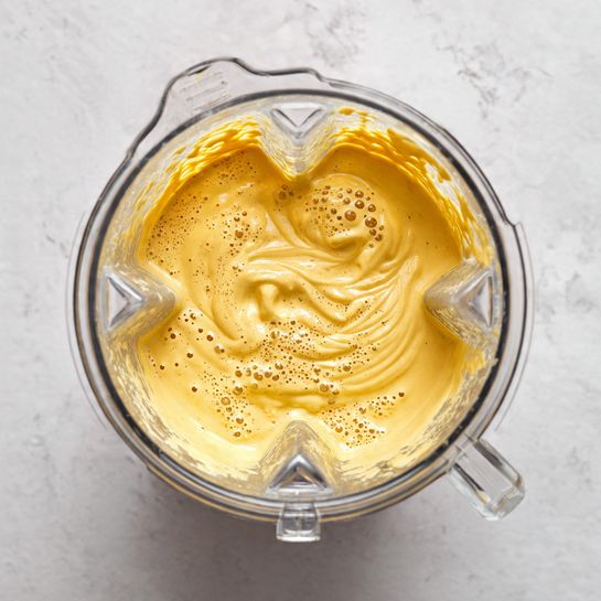 The image shows a thick, creamy mustard yellow mixture inside a clear blender container. The texture appears smooth with some small bubbles and slight swirls on the surface, indicating it has just been blended. The container has four small clear grips around the sides, and the background is a white marbled texture. photo taken with an iphone --ar 4:5 --v 7