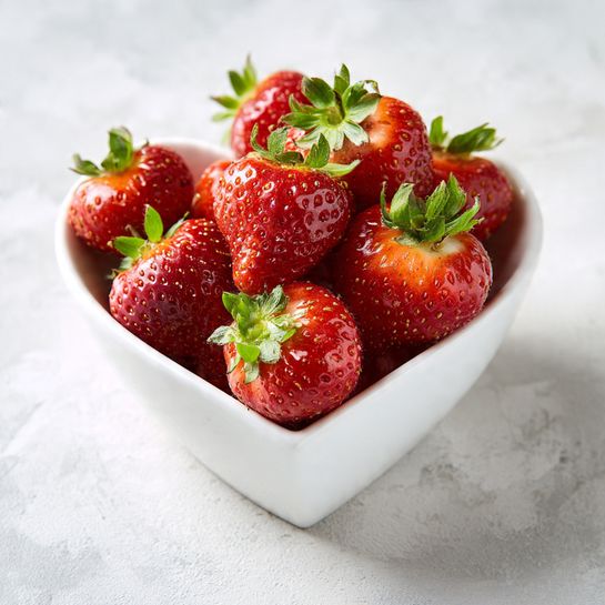 A white heart-shaped bowl is filled with fresh, bright red strawberries with green leafy tops, some strawberries spilling out slightly onto a white marbled surface. The strawberries are shiny and have a smooth texture, with small seeds visible on their skin. The scene is simple, focusing on the vivid red of the berries contrasted with the white bowl and white marbled background. Photo taken with an iphone --ar 4:5 --v 7