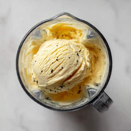 A clear blender jar on a white marbled surface holds a scoop of pale yellow ice cream with several small black vanilla bean specks on top, showing a creamy texture and soft fold patterns, viewed from above. photo taken with an iphone --ar 4:5 --v 7