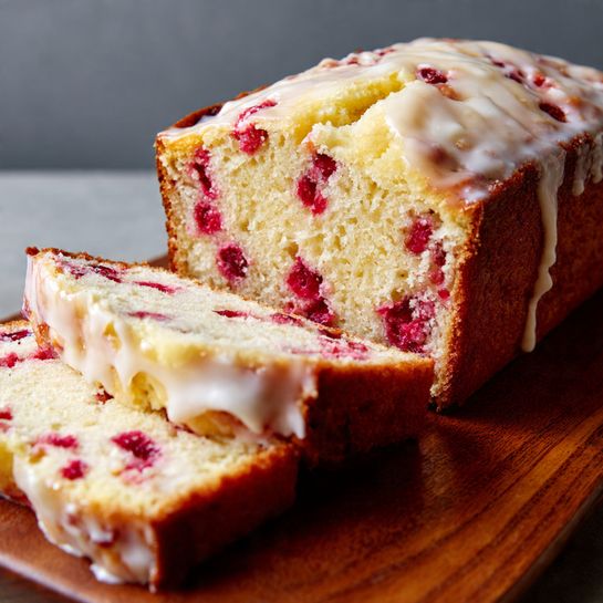 A loaf of cake sits on a wooden board with two thick slices placed in front, showing a soft, light yellow crumb filled with bright red berries evenly spread throughout. The top and the sliced surfaces are covered with a smooth, creamy white glaze that slightly melts and drips over the edges. The top crust is golden brown and slightly cracked, contrasting with the moist texture inside. The whole cake looks rich and inviting with a mix of creamy and fruity textures. Photo taken with an iphone --ar 4:5 --v 7