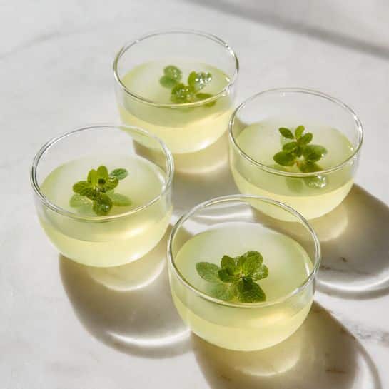 Four clear glass cups are filled with pale yellow liquid, each topped with a small fresh green mint leaf floating on the surface. The cups are arranged closely together on a white marbled surface with soft shadows falling behind them. The liquid inside each cup is smooth and uniform in color. photo taken with an iphone --ar 4:5 --v 7