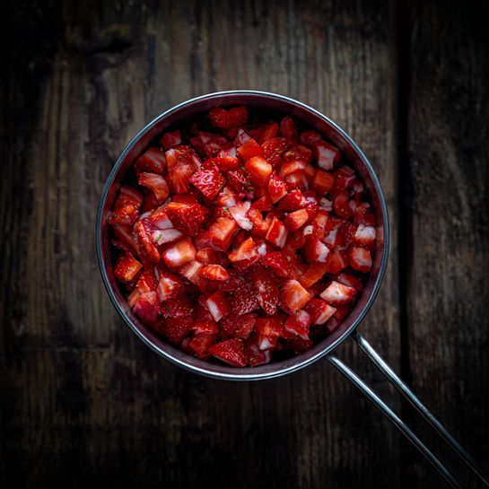 A metal measuring cup filled to the top with small, irregular pieces of bright red and white chopped strawberries. The strawberries show a mix of their red outer skin and white inner flesh, creating a contrast in the cup. The cup has two thin metal handles extending out to the right. The background is a dark wood texture. photo taken with an iphone --ar 4:5 --v 7