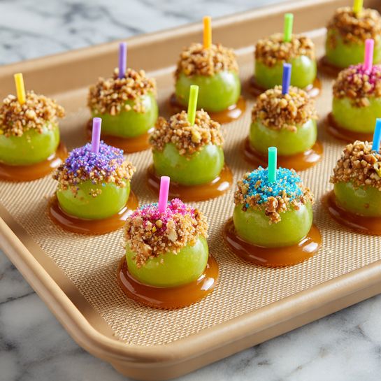The image shows a baking tray filled with small green grapes, each dipped halfway in light brown caramel and rolled in crushed nuts. Each grape is held upright by a colorful toothpick in shades of pink, blue, yellow, green, and orange. The caramel creates a shiny smooth layer around the bottom part of each grape, while the crushed nuts add a rough, textured layer on the caramel. The grapes are neatly arranged in rows on a beige silicone baking mat. The background surface is a white marbled texture. photo taken with an iphone --ar 4:5 --v 7