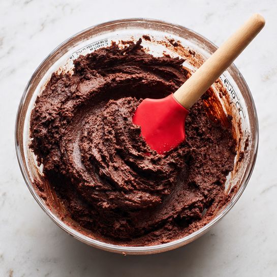 A clear glass bowl filled with dark brown, thick chocolate dough sits on a white marbled surface. The dough is textured with soft peaks and swirls, showing a slightly crumbly but moist consistency. A red spatula with a light wooden handle is pushed into the dough, with part of the dough sticking to it, creating natural folds. The bowl has measurement markings visible on its inner side. The lighting highlights the rich chocolate color and the matte texture of the dough. photo taken with an iphone --ar 4:5 --v 7
