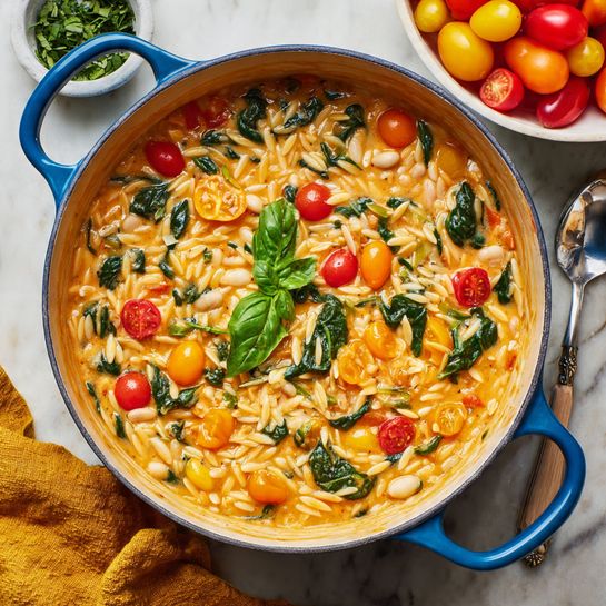 A round blue cooking pot filled with creamy orange orzo pasta mixed with green spinach leaves and sliced colorful cherry tomatoes in red, yellow, and orange. The orzo looks soft and slightly thick with visible spoon-sized pieces of white beans. In the center, there is a fresh bright green basil leaf garnish. The pot sits on a white marbled surface next to a white bowl filled with more colorful cherry tomatoes and a mustard yellow cloth nearby. Photo taken with an iphone --ar 4:5 --v 7